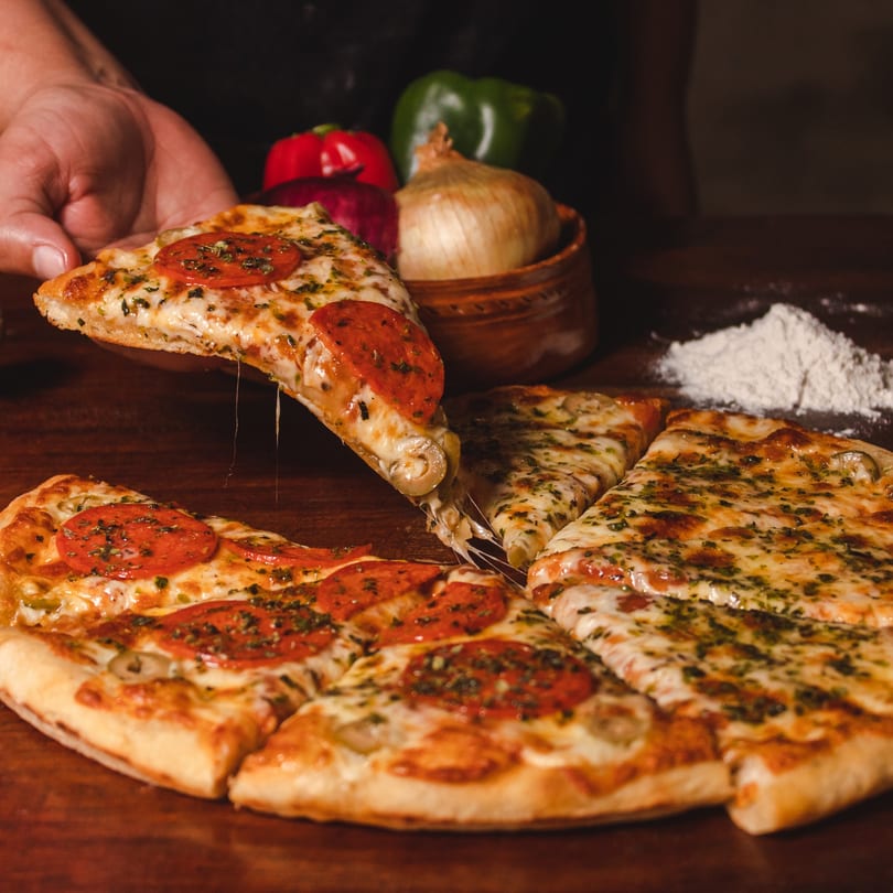 Hand lifting a slice of pepperoni pizza with melted cheese, alongside other pizza slices and ingredients.